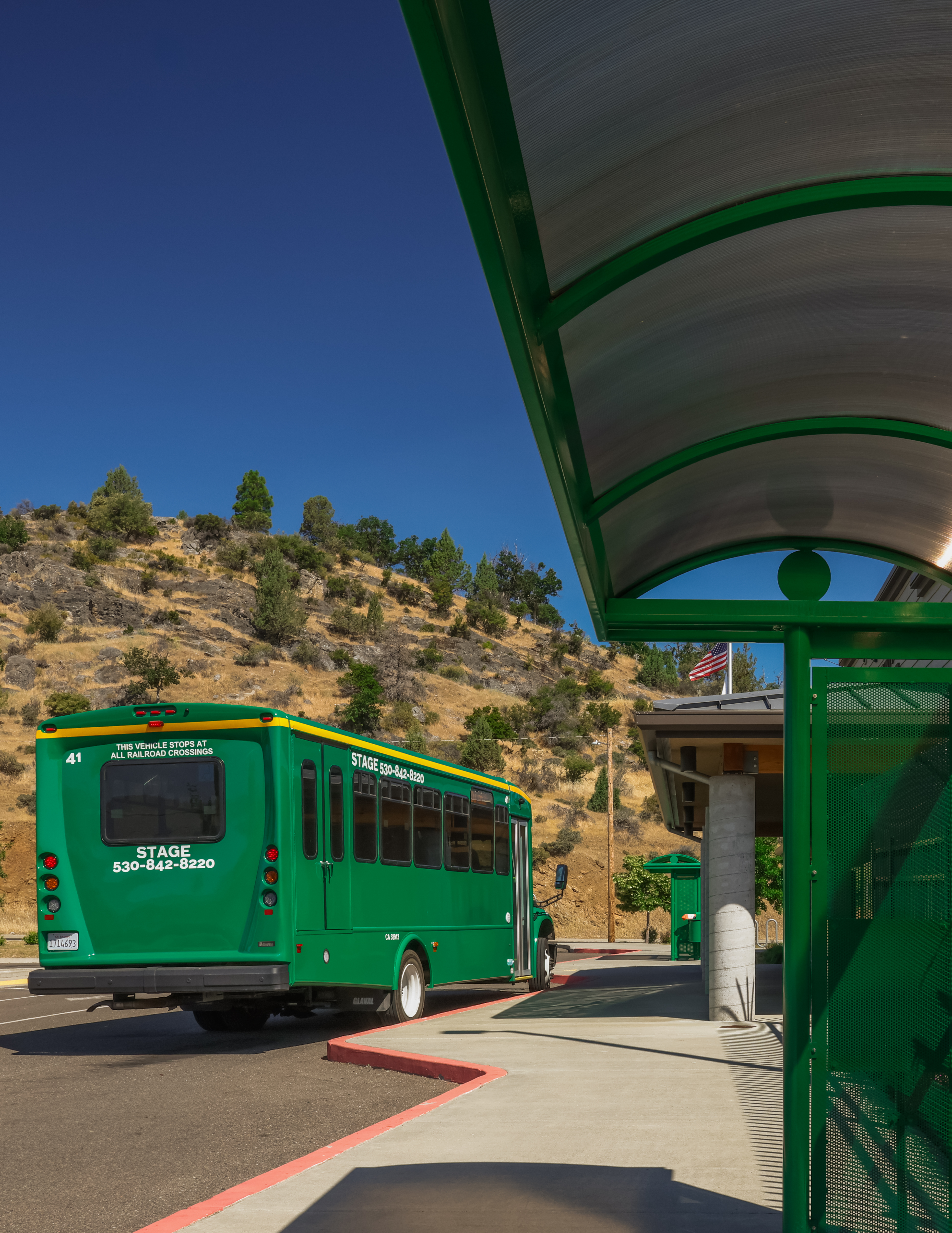 Green public transit bus at a covered stop, beside dry California hills under a bright blue sky; the scene feels sunny and calm.