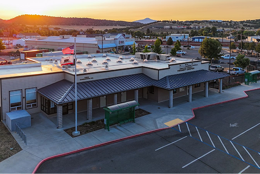 A single-story public building with an American flag, parking lot, and surrounding businesses, photographed at sunset with hills in the background.
