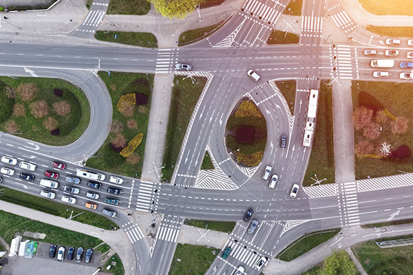 Aerial view of a busy multi-lane intersection with cars, buses, and marked pedestrian crosswalks, surrounded by green spaces and sidewalks.