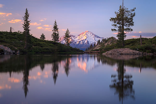 A calm lake reflects pine trees and a snow-capped mountain at sunset, with soft clouds in the sky and green vegetation along the shore.