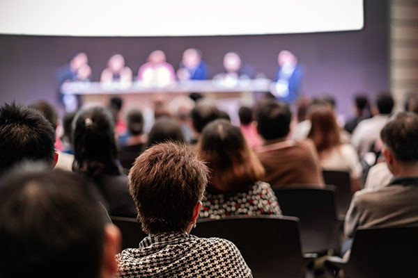 Audience members seated and listening to a panel of speakers at a conference or seminar, with the speakers blurred in the background.