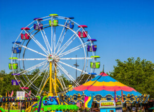 Colorful carnival under a clear blue sky, featuring a large Ferris wheel, bright ride tents, leafy trees, and crowds enjoying a lively fair.