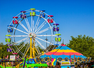 Colorful carnival under a clear blue sky, featuring a large Ferris wheel, bright ride tents, leafy trees, and crowds enjoying a lively fair.